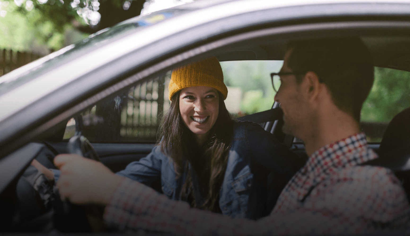 Une passagère souriante en bonnet jaune et un conducteur en chemise à carreaux échangent un moment convivial en voiture.
